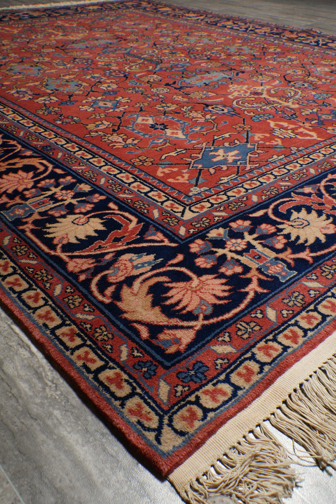 A close-up of a traditional oriental rug with a red field, navy blue borders, and cream floral motifs, showing the corner with beige fringe.