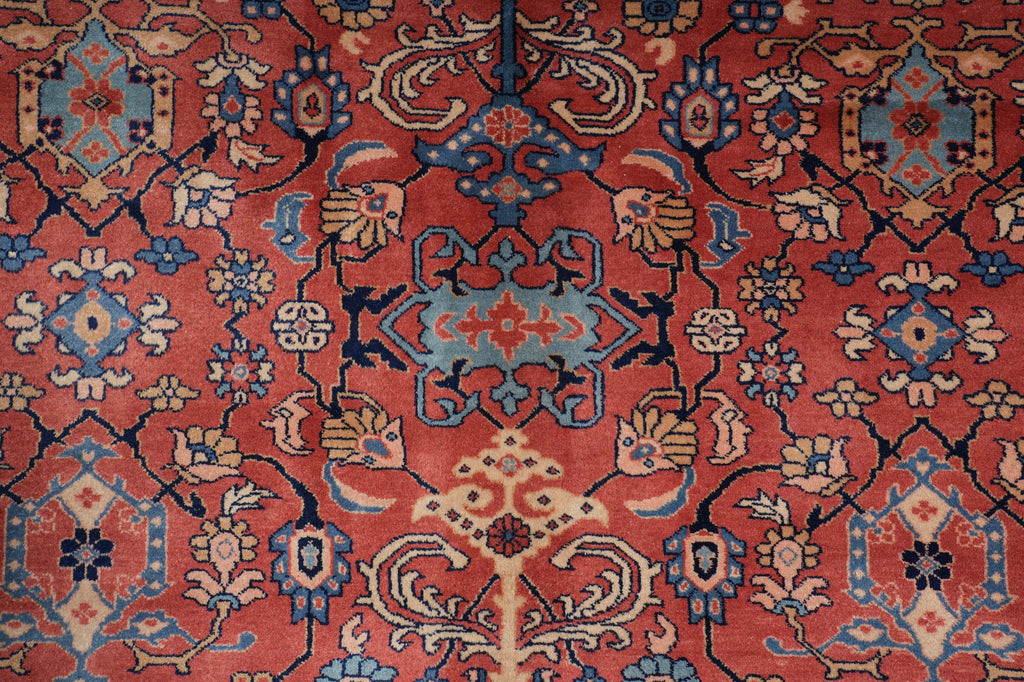 A close-up of a traditional patterned rug with a coral-red field, blue and beige floral motifs, and dark navy outlines.