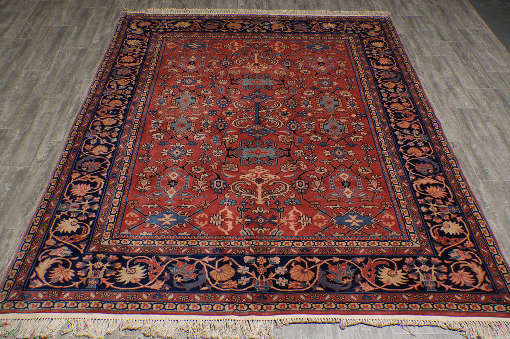 A traditional oriental area rug with a complex floral and geometric pattern, featuring a dominant red field, dark navy borders, and accents of beige and blue, laid out on a gray wood-look floor.