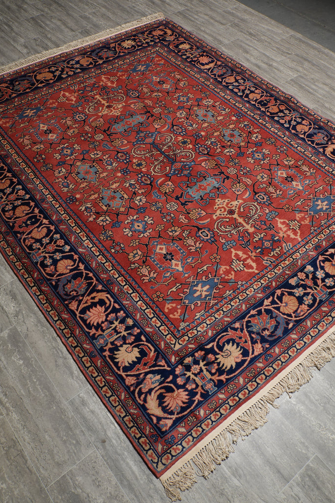 A traditional Persian-style area rug with a red field, navy blue borders, and beige and blue floral motifs, shown laid out on a gray tiled floor.