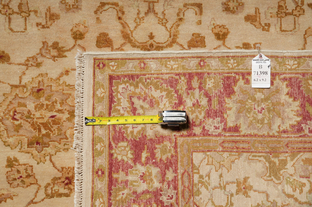 A close-up of a patterned area rug corner with a yellow tape measure laid across the fringe and a small product tag attached, showing the rug's beige, pink, and gold design.