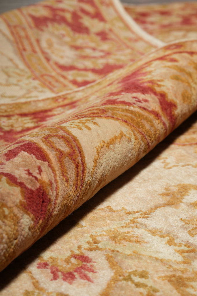 A close-up of a patterned fabric or rug with a cream base and swirling red, rust, and gold motifs, shown folded to reveal texture and pattern detail.