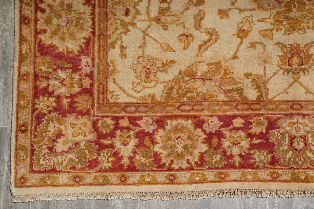 A close-up of a traditional patterned rug corner, showing a cream field with floral motifs and a deep red border with gold and beige detailing.