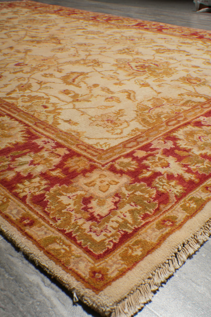 A close-up of a patterned area rug corner showing cream, rust red, and gold floral motifs with fringed edges.