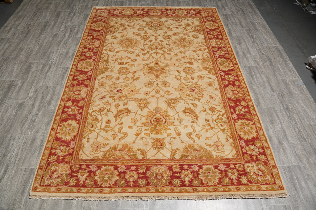 A rectangular traditional area rug with a cream center and red borders, featuring intricate floral and vine patterns in gold and red tones, laid out on a gray wood-look floor.
