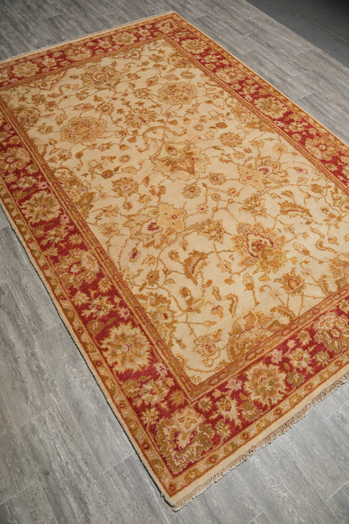 A rectangular traditional area rug with an ornate floral pattern, featuring a cream center field and deep red borders, laid out diagonally on a gray wood-look floor.