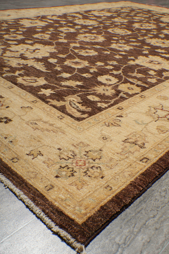 A close-up of a traditional patterned area rug showing its corner and fringe, featuring a dark brown central field with beige floral motifs and a wide beige border on a gray floor.