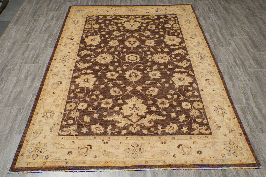 A rectangular area rug with an ornate floral pattern, featuring a dark brown center field surrounded by beige and brown borders, laid out on a gray wood-look floor.