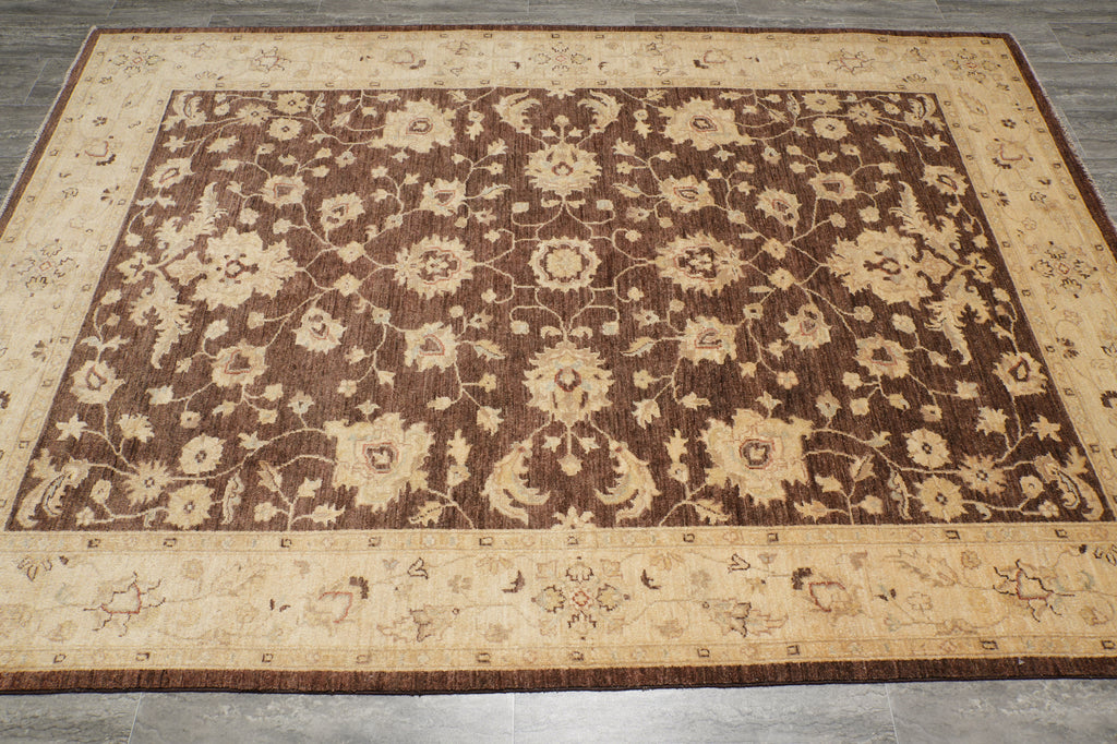 A rectangular area rug with an ornate floral and vine pattern, featuring a dark brown center field and a beige border, laid out on a gray tiled floor.