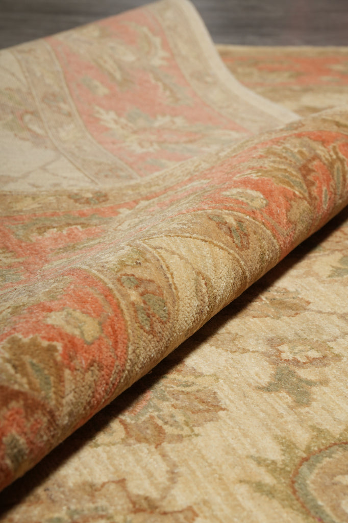 A close-up of a patterned rug with a rolled section showing its texture and design. The rug features a traditional floral or paisley motif in muted tones of beige, dusty pink, ochre, and olive green.