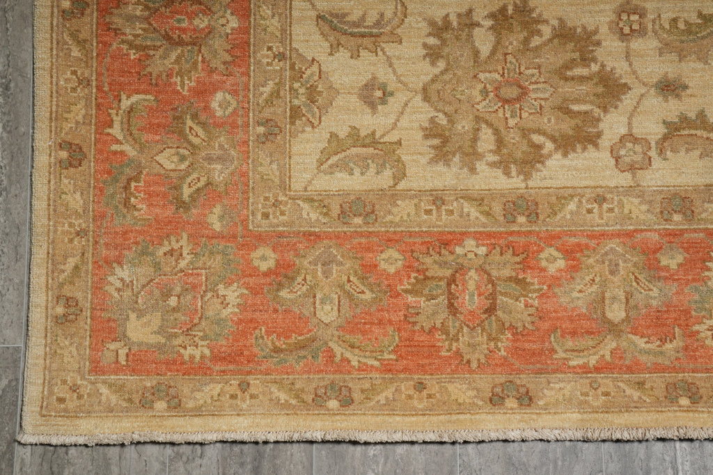 A close-up of a traditional patterned rug corner, showing a warm rust-red field, beige border, and muted green and tan floral motifs on a gray floor.