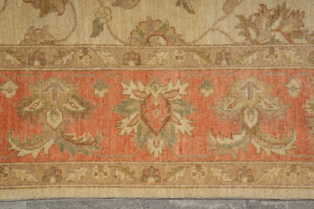 A close-up of a vintage-style area rug featuring a wide central band in muted coral orange with beige and sage green floral motifs, bordered by a beige floral field and patterned edge.