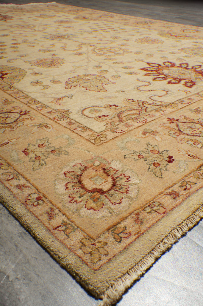 A close-up view of an ornate area rug corner, showing a cream and beige base with red and muted green floral motifs and a fringed edge on a gray floor.