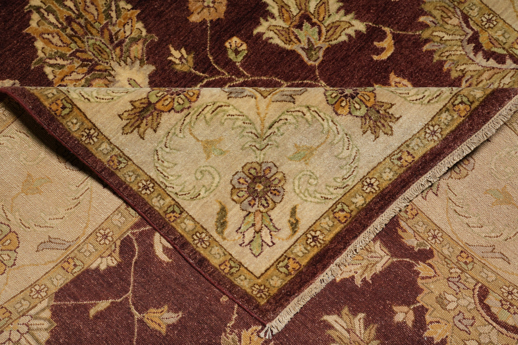 A close-up of a traditional patterned rug showing its corner and fringe detail. The design features a deep burgundy background with a cream-colored central field and floral motifs in beige, gold, and muted green.