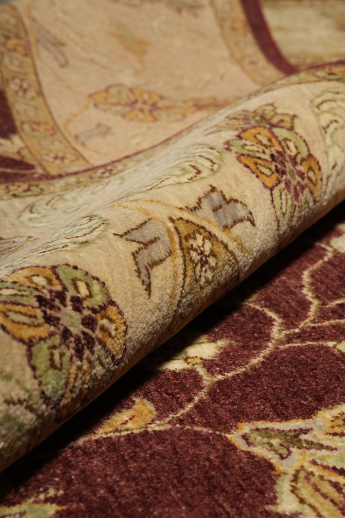 Close-up of a patterned area rug with a rolled section showing its texture and design. The rug features a traditional floral motif in burgundy, beige, ochre yellow, and olive green.