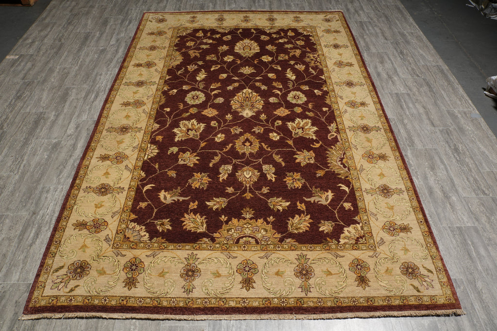 A rectangular area rug with an ornate floral pattern, featuring a dark brown center field and a beige border, laid out on a gray wood-look floor.