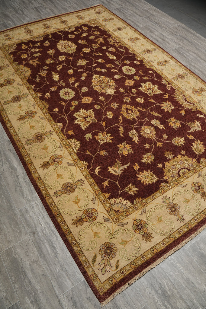 A rectangular area rug with a traditional floral pattern, shown from an elevated angle on a gray wood-look floor. The rug features a deep burgundy central field with beige floral motifs, surrounded by a wide beige border with decorative motifs and a dark burgundy edge.