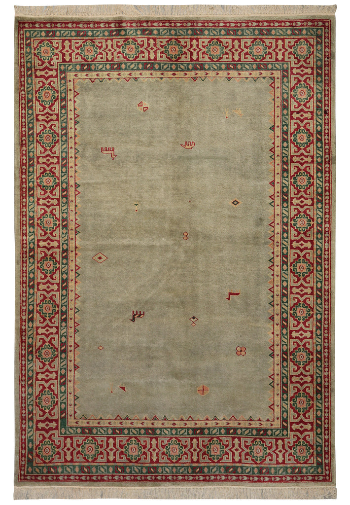A rectangular area rug with a muted sage green center field, a red and green geometric border, and cream fringe on the top and bottom.