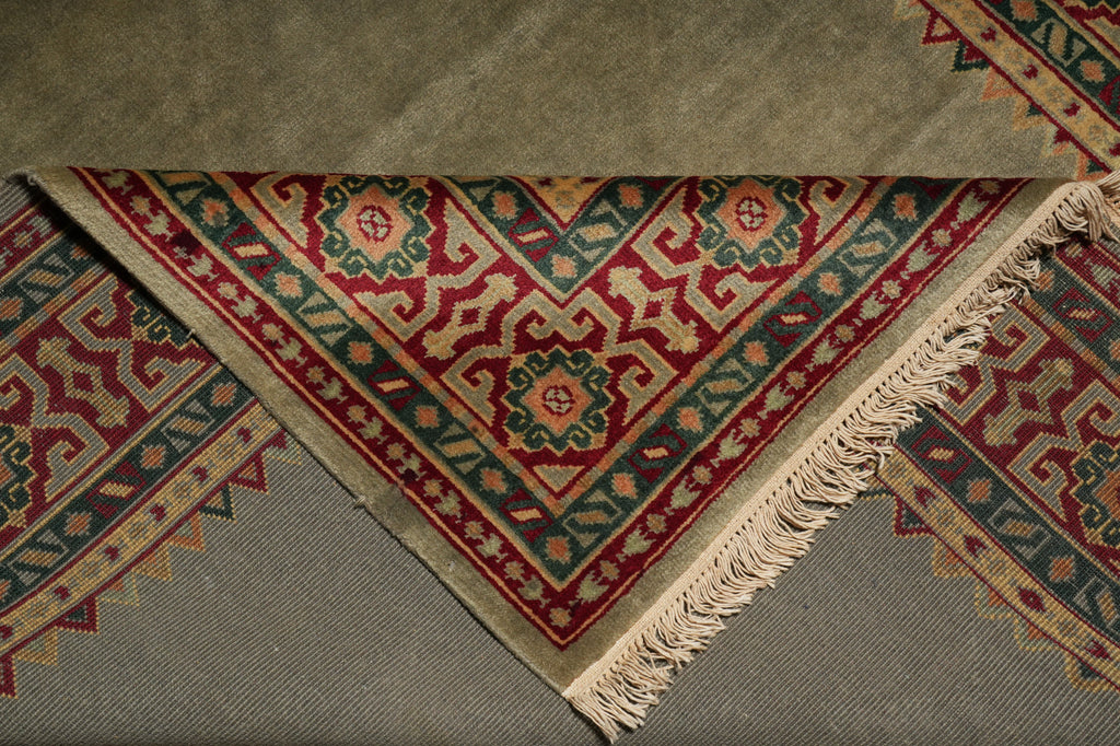 A close-up of a folded patterned rug showing its corner and fringe detail. The rug features a complex geometric design in red, green, beige, and blue, layered over a solid taupe-gray backing.