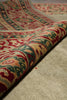 Load image into Gallery viewer, A close-up of a folded patterned rug showing its detailed design and texture. The rug features a complex, traditional pattern in red, green, beige, and cream, draped over a neutral beige textured surface.