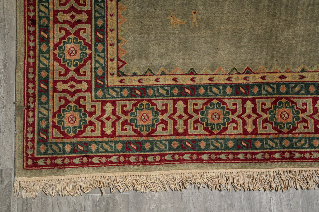 A close-up of a patterned area rug corner with a beige field, red and green geometric borders, and cream fringe along the bottom edge.