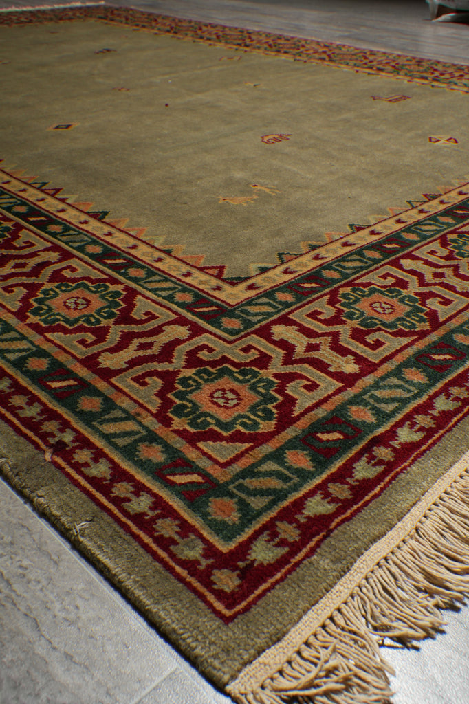 A close-up of a patterned area rug with a beige base, featuring a red, green, and orange geometric border and fringe on one end.