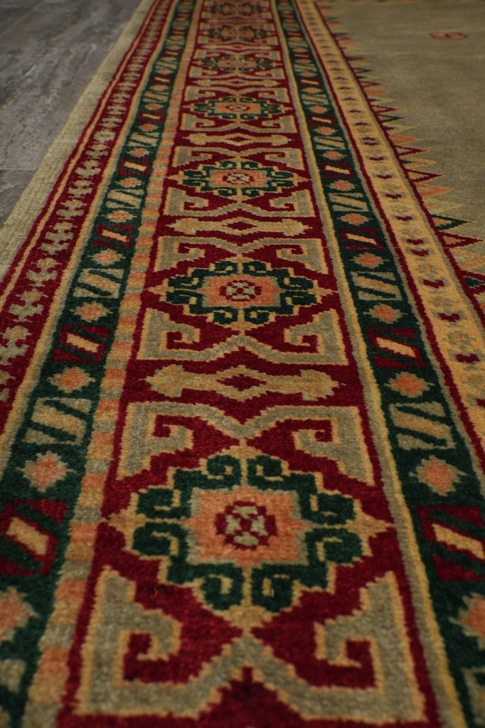 A close-up of a woven geometric rug or textile with repeating ornamental motifs in red, beige, green, and cream, shown against a neutral gray surface.
