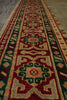 Load image into Gallery viewer, A close-up of a woven geometric rug or textile with repeating ornamental motifs in red, beige, green, and cream, shown against a neutral gray surface.
