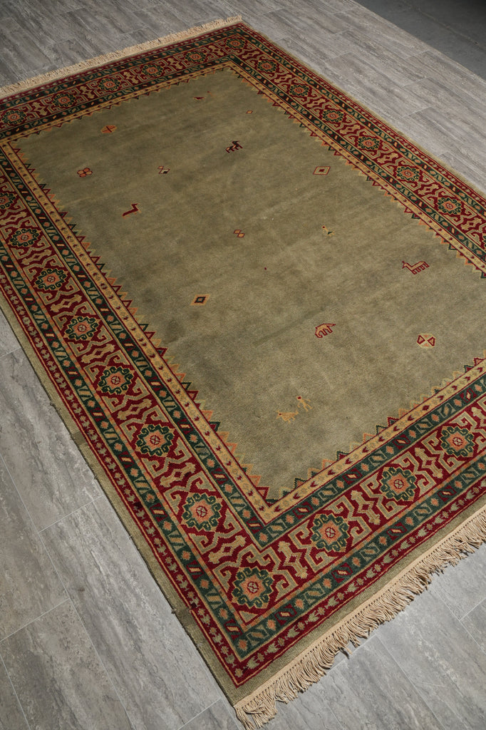 A rectangular area rug with a beige center field and a detailed red, green, and brown geometric border, shown on a gray wood-look floor.