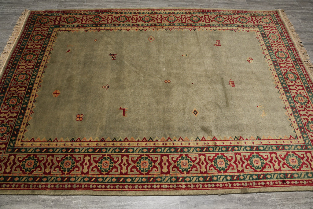 A rectangular area rug with a beige center field and a detailed red, green, and black geometric border, laid flat on a gray floor.