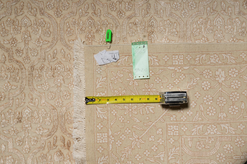 A beige patterned area rug with a fringed edge is shown on a white surface, with a yellow tape measure, small green clips, and handwritten notes placed on top to indicate dimensions.