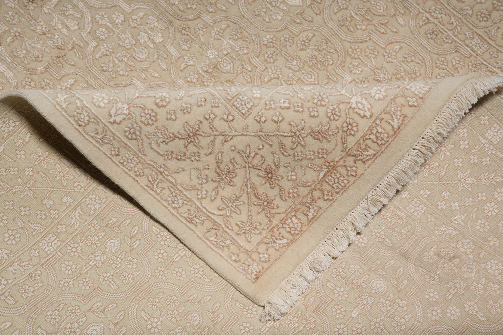A close-up of a folded cream-colored area rug with a detailed floral and geometric pattern and fringed edges, laid over a matching patterned rug.