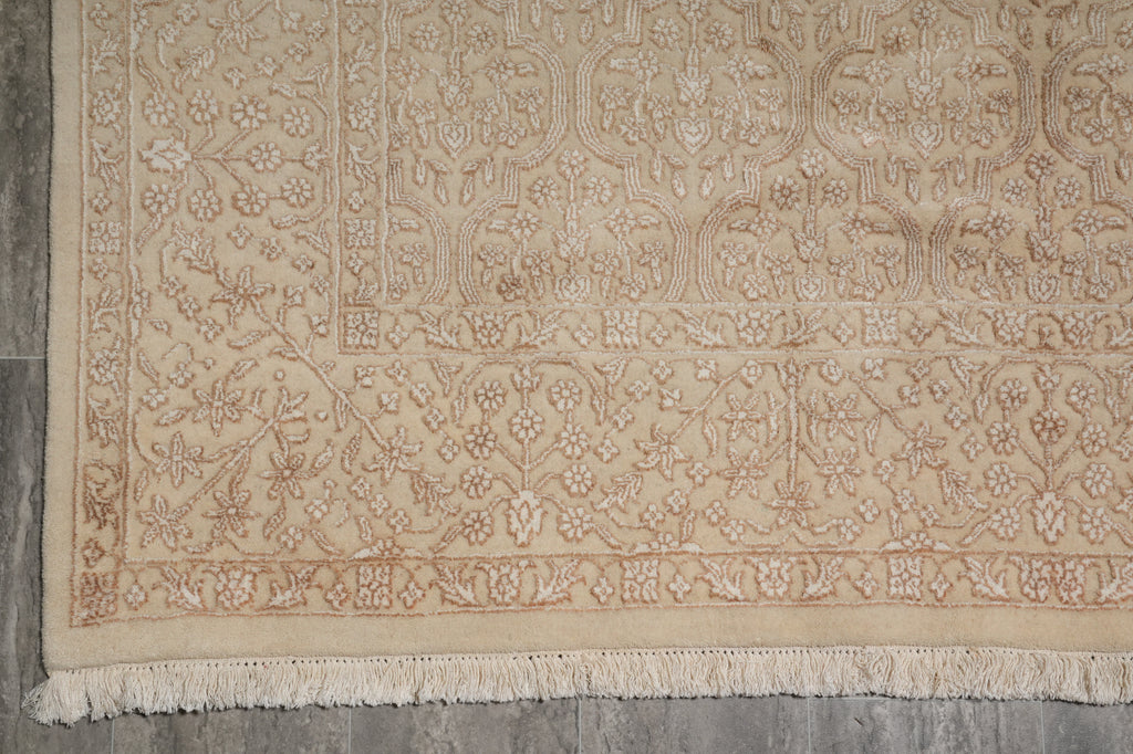 A close-up of a beige area rug with an intricate floral and botanical pattern and fringed edges, laid out on a gray floor.
