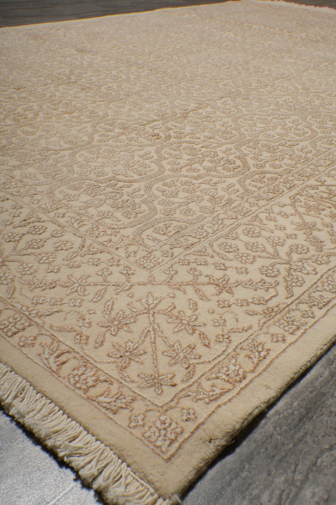 A close-up of a beige area rug with a traditional floral pattern and fringed edge, shown on a neutral gray floor.