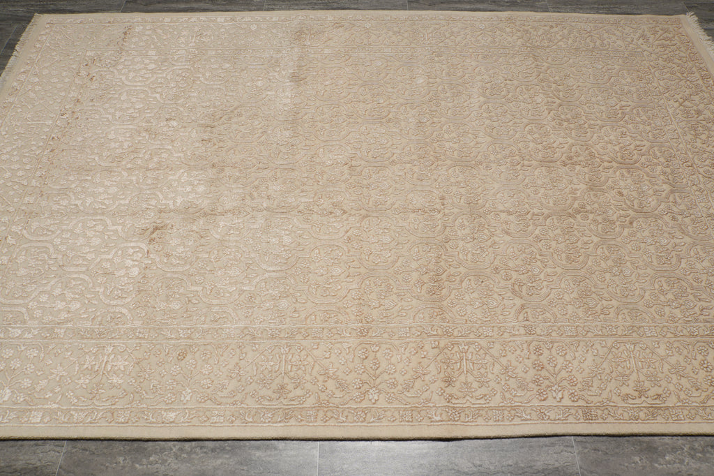 A large rectangular area rug in a light beige or cream color with an intricate, faded traditional floral pattern and a decorative border, laid out on a dark gray tiled floor.