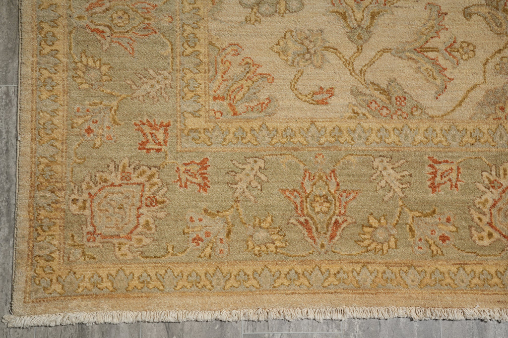 A close-up of an area rug showing its corner and fringe, featuring a traditional floral pattern in beige, muted green, and terracotta orange.
