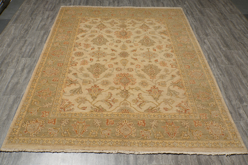 A rectangular area rug with an intricate traditional floral pattern, featuring a beige center, a muted sage green border, and accents of rust orange and brown, laid out on a gray wood-look floor.