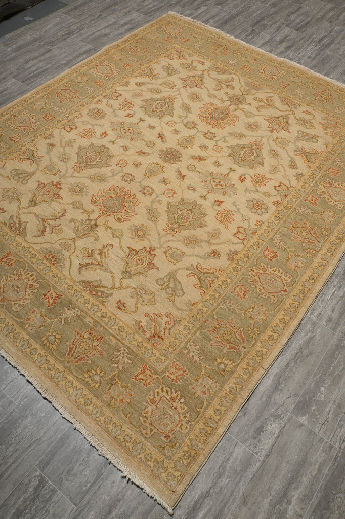A rectangular area rug with an intricate traditional floral pattern, shown laid out on a gray wood-look floor. The rug features a warm beige center, a muted sage green border, and accents of terracotta and rust orange.