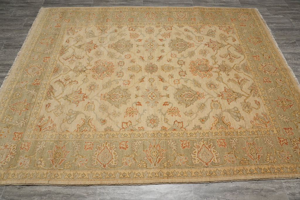 A large rectangular area rug with an intricate traditional floral pattern, laid out on a dark gray tiled floor. The rug features a warm beige center field with a wide border in muted sage green and terracotta orange.