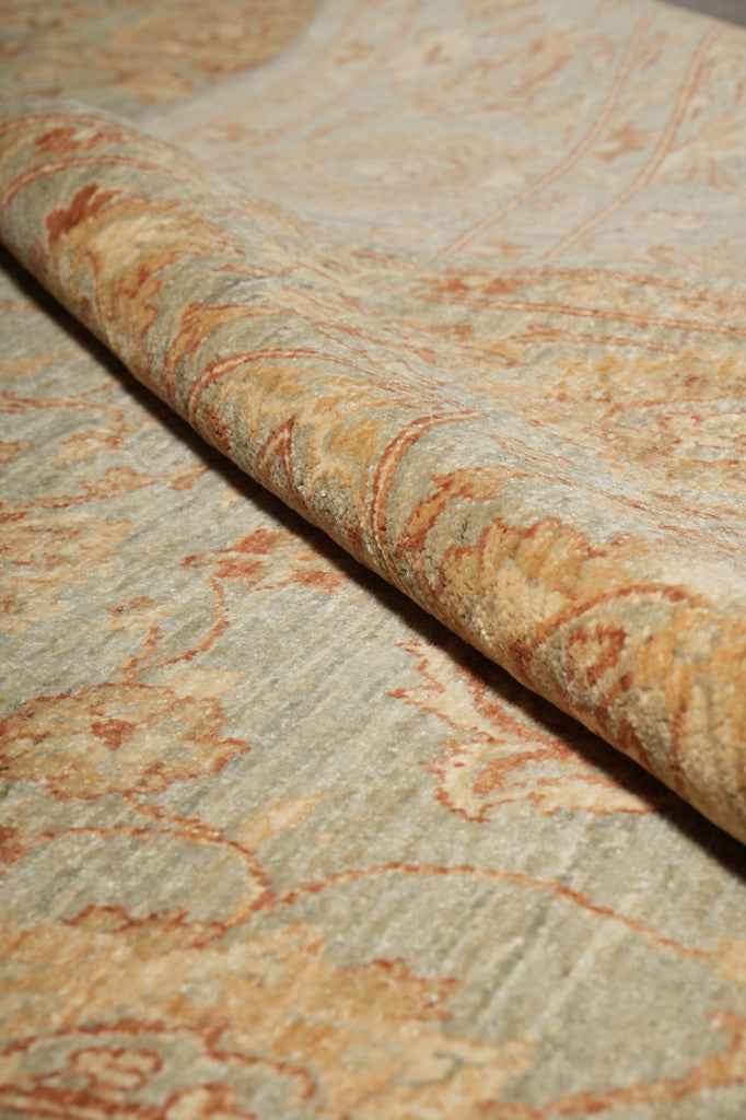 A close-up of patterned upholstery fabric with a rolled section showing the texture and color. The material has a muted sage green base with a warm beige and rust-orange floral damask pattern.