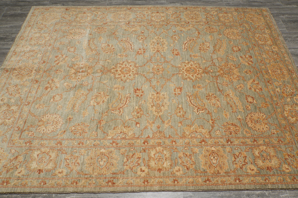 A rectangular area rug with an intricate traditional floral and paisley pattern in muted beige, tan, and rust-orange tones over a dusty sage green background, shown flat on a gray wood floor.