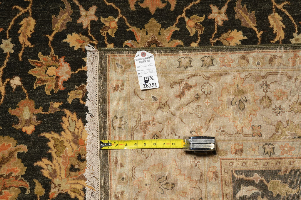 A close-up of a patterned area rug with a beige center and dark floral border, shown with a yellow tape measure and a white tag on a dark rug background.