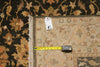 Load image into Gallery viewer, A close-up of a patterned area rug with a beige center and dark floral border, shown with a yellow tape measure and a white tag on a dark rug background.