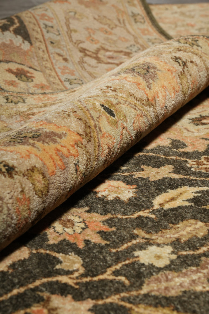 A close-up of a patterned rug with a rolled section showing the underside, featuring a detailed floral and scroll design in beige, brown, black, and muted orange tones.