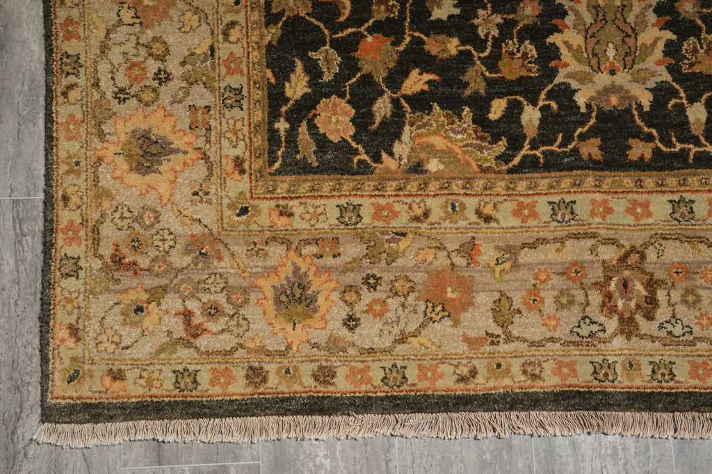 A close-up of an ornate area rug corner showing detailed floral motifs, traditional borders, and fringe, laid out on a gray wood floor.