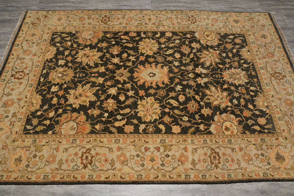 A rectangular area rug with an ornate floral pattern, featuring a dark central field surrounded by a beige border, laid out on a gray tiled floor.