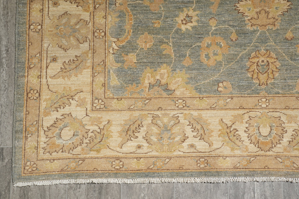 A close-up of a traditional patterned rug corner, showing a dusty blue field with beige and ochre floral motifs and a cream border, laid on a gray floor.