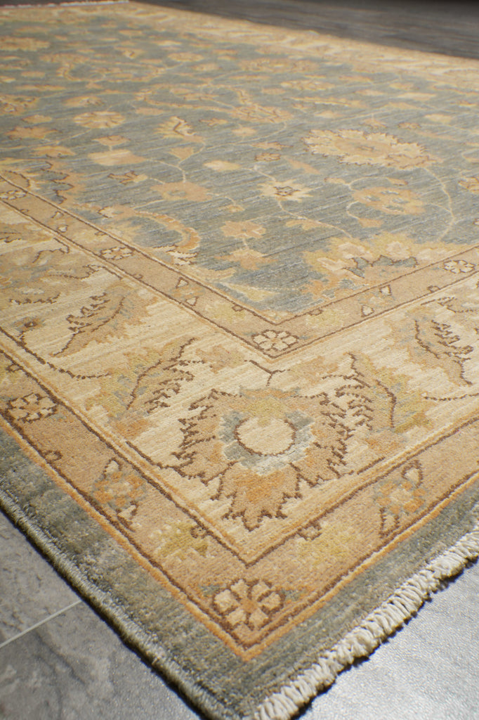 A close-up view of an area rug showing its corner detail, featuring a traditional floral pattern in beige, cream, and muted blue-gray tones with a fringed edge.