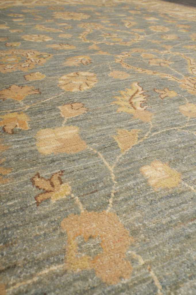 A close-up of a textured fabric or rug with a muted blue-gray base and a repeating floral pattern in tan and beige.
