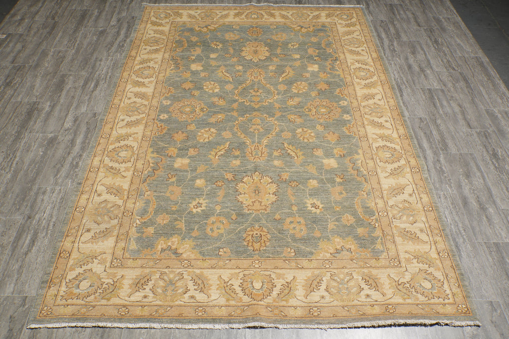 A rectangular area rug with an ornate traditional floral pattern, featuring a muted blue-gray center field and beige/gold borders, laid out on a gray wood-look floor.